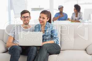 Couple using laptop with colleagues in background at creative of