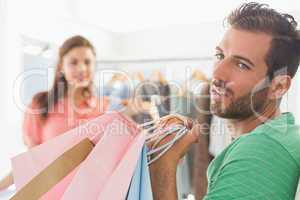 Bored man with shopping bags while woman by clothes rack