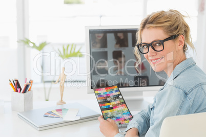 Blonde designer holding colour chart at her desk smiling at came