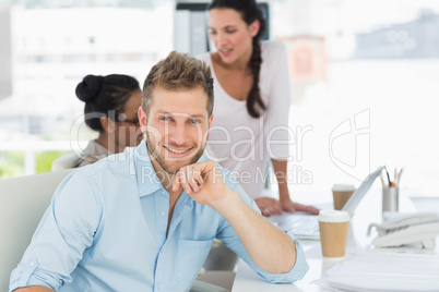 Handsome man smiling at camera while colleagues talk at desk