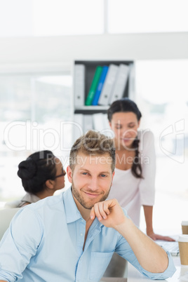 Handsome man smiling at camera while colleagues chat at desk