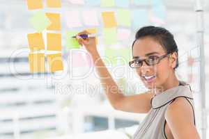 Smiling designer writing on sticky notes on window looking at ca