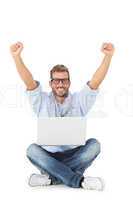 Handsome young man cheering at camera with laptop sitting on flo