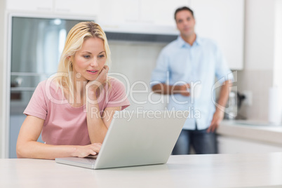 Woman using laptop with man drinking coffee in kitchen