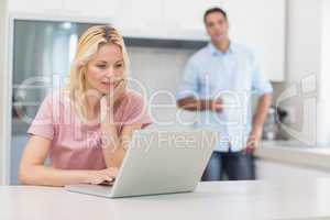Woman using laptop with man drinking coffee in kitchen