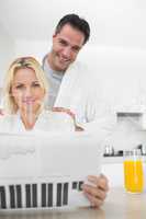 Smiling couple in bathrobes reading newspaper in kitchen