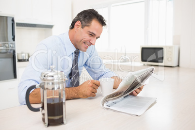Smiling well dressed man with coffee cup reading newspaper in ki