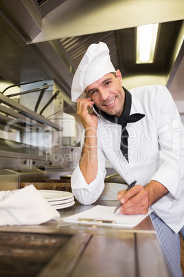 Male cook writing on clipboard while using cellphone in kitchen