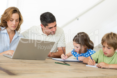 Mother using laptop while father assisting children in coloring