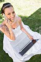 Happy bride with laptop using cellphone on grass