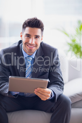 Businessman sitting on sofa using his tablet pc smiling at camer