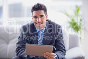 Businessman sitting on sofa using his tablet smiling at camera