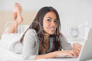 Pretty girl lying on bed using her laptop smiling at camera