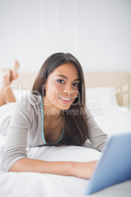 Pretty girl lying on bed using her tablet smiling at camera