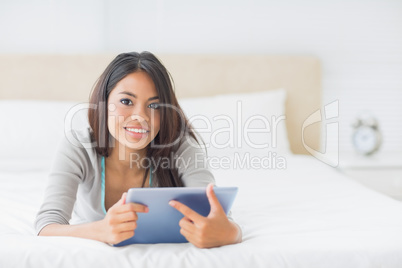 Young pretty girl lying on her bed using her tablet smiling at c