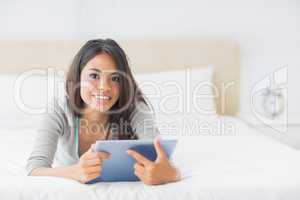 Young pretty girl lying on her bed using her tablet smiling at c