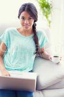 Pretty girl sitting on a sofa using laptop smiling at camera