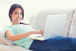 Pretty girl lying on sofa using her laptop smiling at camera