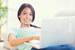 Happy girl lying on sofa using her laptop smiling at camera