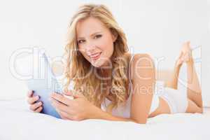 Pretty young blonde smiling at camera on bed using tablet