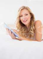 Cute young blonde smiling at camera on bed using tablet