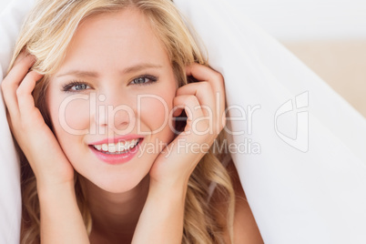 Cheerful blonde smiling at camera from under the duvet