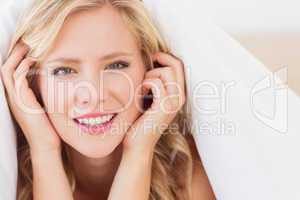 Cheerful blonde smiling at camera from under the duvet