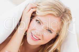 Pretty young blonde smiling at camera from under the duvet