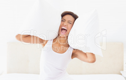 Screaming brunette sitting on bed covering ears with pillow