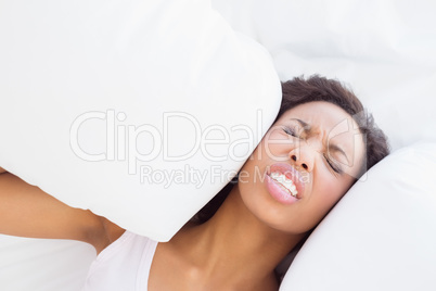 Stressed brunette sitting on bed covering ears with pillow