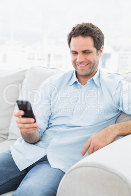 Cheerful man sitting on the couch sending a text with smartphone