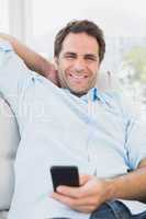 Smiling man sitting on the sofa texting on his phone looking at