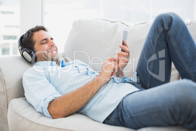 Happy handsome man lying on sofa using tablet and listening to m