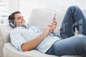 Happy handsome man lying on sofa using tablet and listening to m