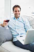 Handsome man relaxing on sofa with glass of red wine using lapto