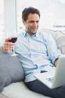 Happy man relaxing on sofa with glass of red wine using laptop