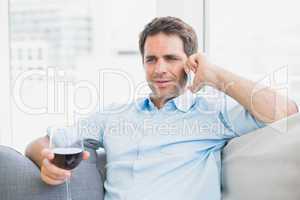 Cheerful man relaxing on sofa with glass of red wine talking on