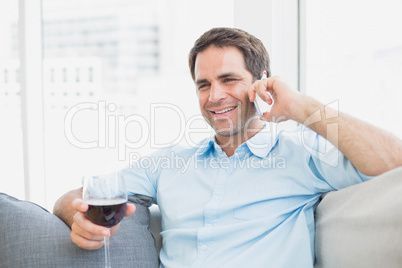 Happy man relaxing on sofa with glass of red wine talking on pho