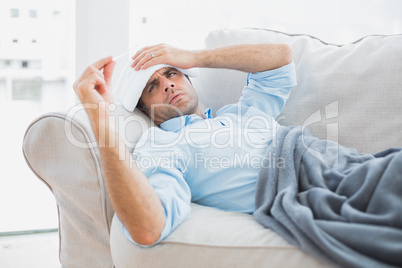 Sick man lying on sofa checking his temperature under a blanket