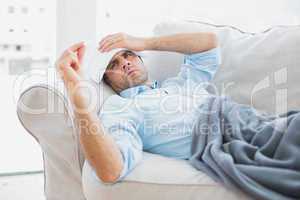 Sick man lying on sofa checking his temperature under a blanket