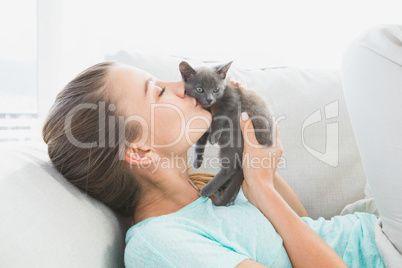 Cheerful woman lying on sofa kissing a grey kitten