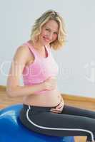 Smiling blonde pregnant woman touching belly on exercise ball