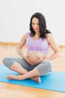 Pregnant brunette sitting on mat in touching her belly