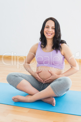 Pregnant brunette sitting on mat in touching her belly smiling a