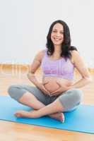 Pregnant brunette sitting on mat in touching her belly smiling a