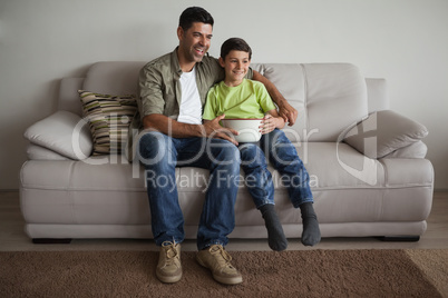 Father and son with popcorn bowl watching tv in living room