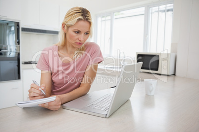 Woman using laptop while writing notes in kitchen