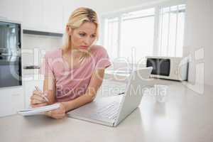 Woman using laptop while writing notes in kitchen