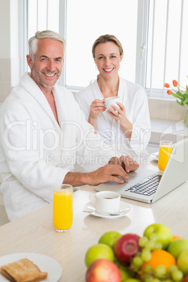 Smiling couple using laptop at breakfast in bathrobes