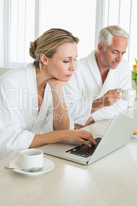 Couple having coffee at breakfast in bathrobes using laptop